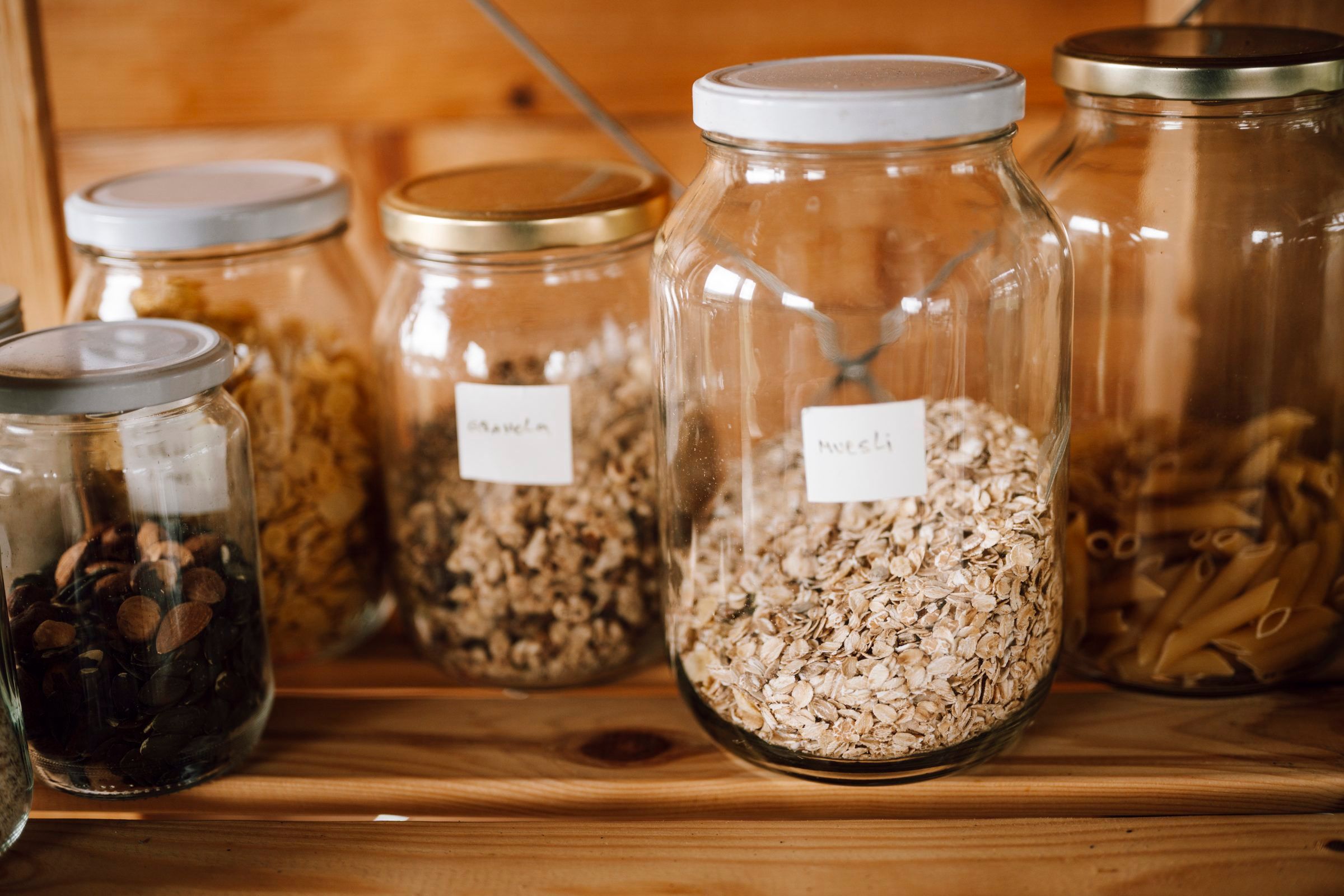 Glass jars of oats, granola, pasta and dried goods with screw-top lids on a wooden shelf, two with small handwritten paper labels.